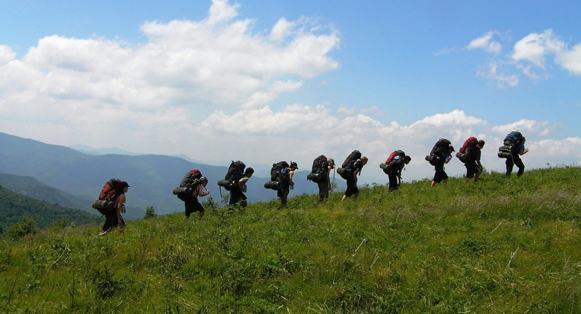 A line of people wearing backpacks hike in a line across a grassy meadow under blue skies. There are mountains in the background. 